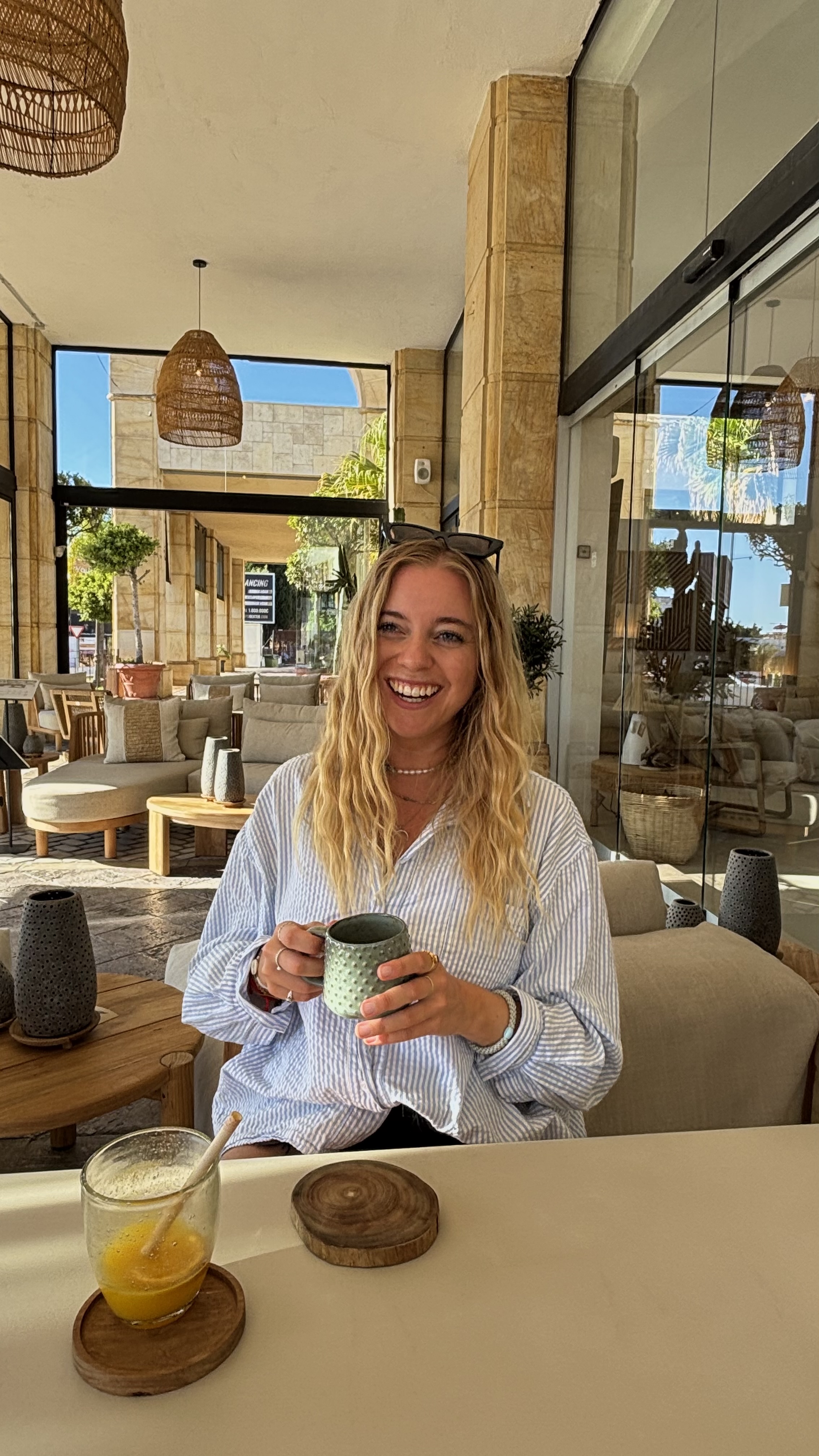 Lächelnde Frau mit langen blonden Haaren an einem Tisch, eine grüne strukturierte Tasse in der Hand, davor ein Glas Orangensaft.